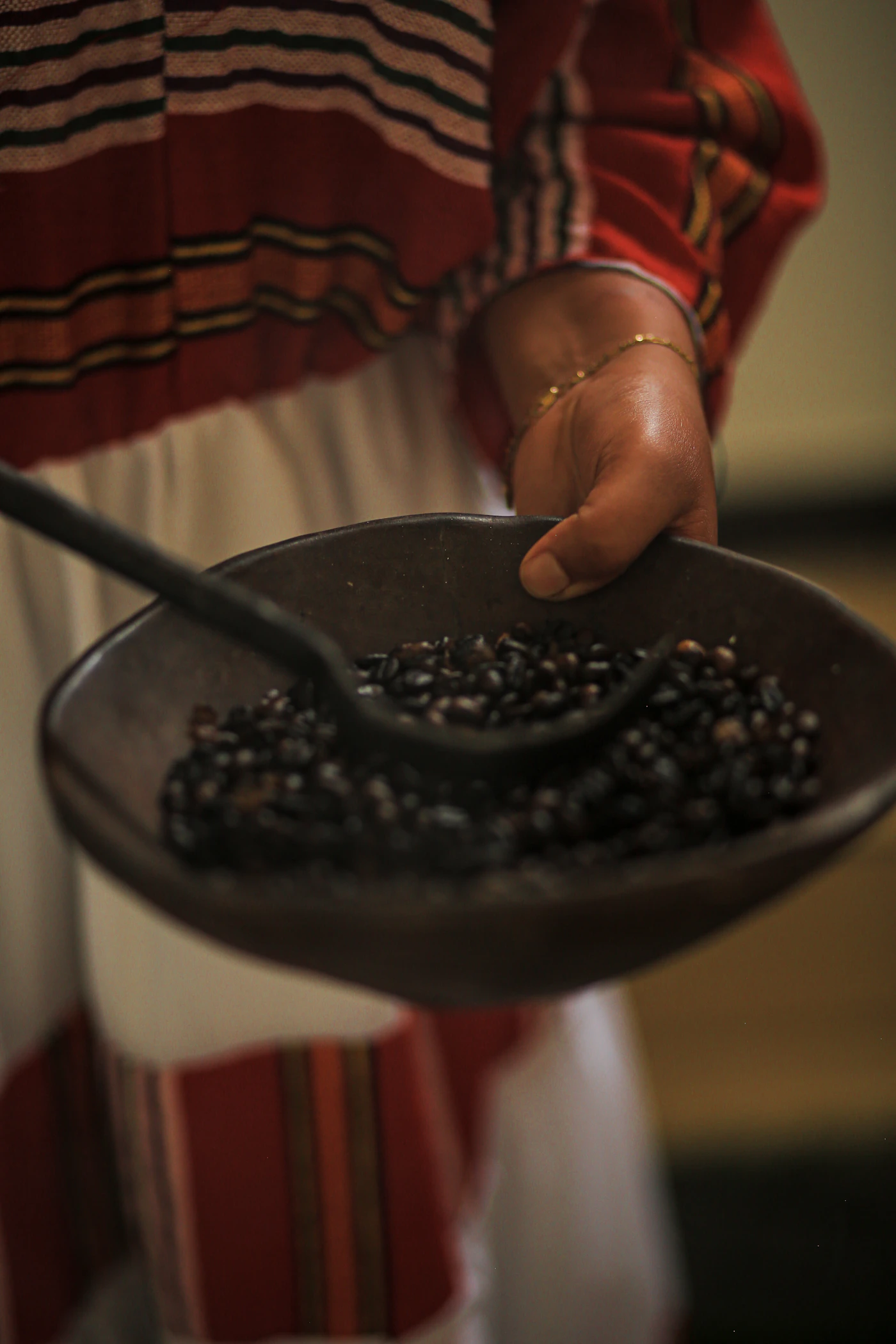 A hand holds a dark pan of freshly roasted coffee beans with a stirring rod, a striped white dress behind