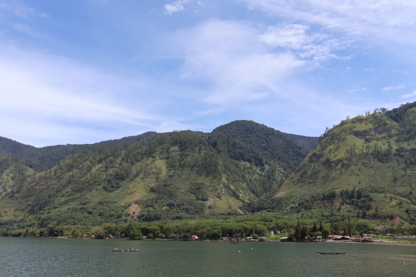 A lake with small boats below steep green hills and a line of lakeside houses under a blue sky